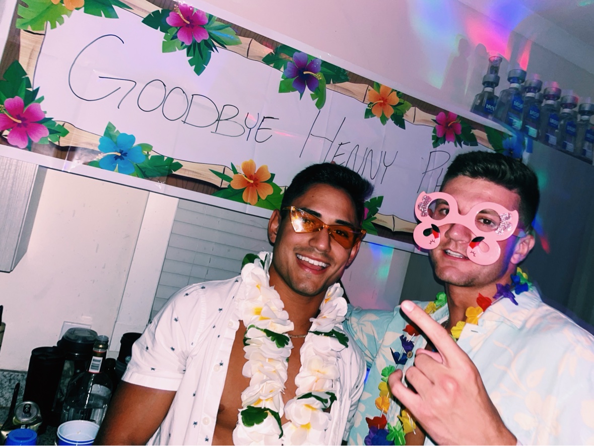 Matthew at a fraternity party in Tampa with a friend, wearing leis in front of a party banner
