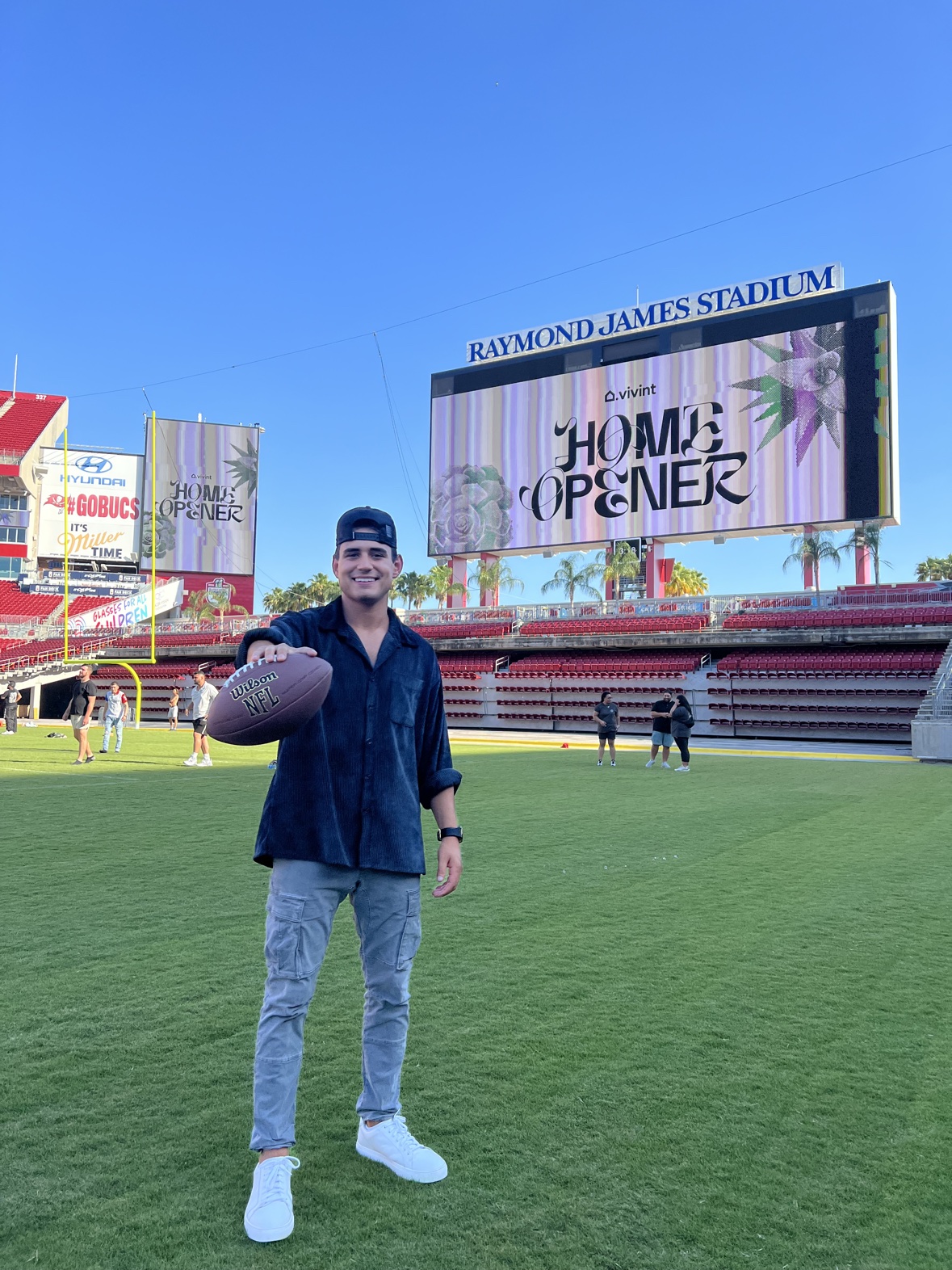Matthew Tenney on the field at Raymond James Stadium holding a football, Vivint Home Opener on the big screen